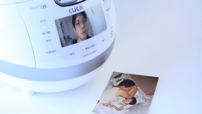 A white Cuckoo brand rice cooker sits on a white counter next to a family photo of a parent alseep next to a young child, their arms intertwined. On the screen of the rice cooker is the face of South Korean theatremaker Jaha Koo.