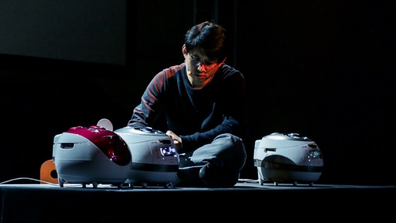 South Korean theatremaker Jaha Koo, an Asian man with short dark brown hair and round glasses, wears a black shirt and sits on top of a table next to three Cuckoo brand rice cookers. His face is lit by a reddish-orange light.