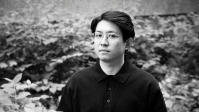 South Korean theatremaker Jaha Koo, a young Asian man with short dark hair and round glasses, wears a black polo shirt and looks towards the camera, greenery behind him, in this black and white image.