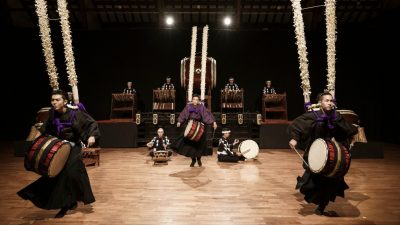 Members of Japanese drumming group Kodo wear traditional attire and play drums of various sizes on stage.