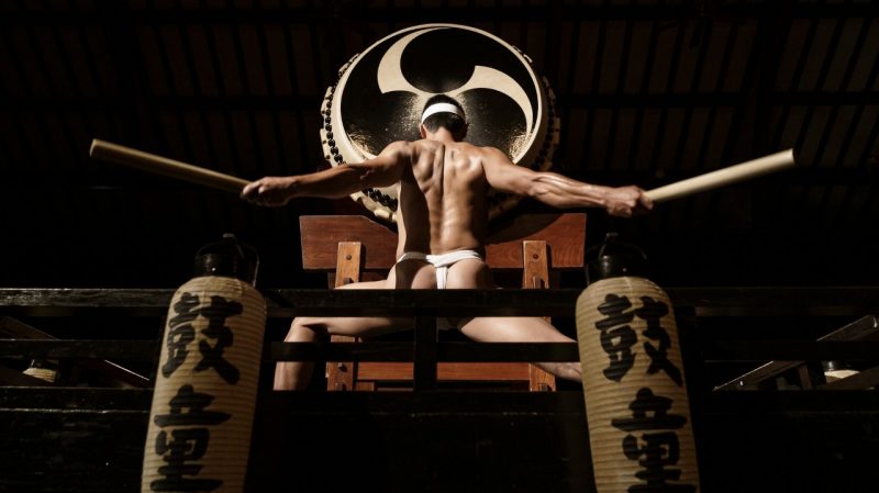  A Japanese drummer wears a white headband and a white fundoshi and extends both arms out side to side, a drum stick in each hand and back muscles all tensed. In front of him is an enormous drum.