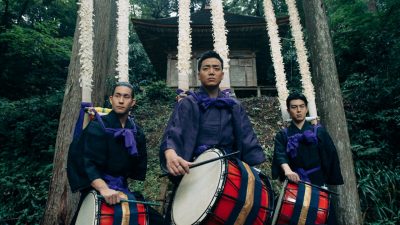 Three members of Japanese drumming group Kodo wear traditional attire and hold a large red drum each. In the background is a forest of trees and a traditional Japanese building.