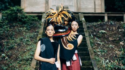 Two members of Japanese drumming group Kodo, two women, wear traditional attire and stand on the steep concrete steps leading to a building in a Japanese forest. The woman at back holds a large dragon mask on her shoulder.