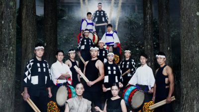 Members of Japanese drumming group Kodo wear traditional attire and hold their instruments on a set of stairs leading to a building in a Japanese forest.