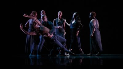 Members of A.I.M by Kyle Abraham wear all black and perform onstage. They group together, one man falling back gracefully as another dancer holds him up and the rest of the dancers look on.