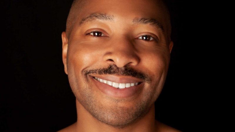 Company artistic director Kyle Abraham, of A.I.M by Kyle Abraham, smiles towards the camera against a black background in this headshot. Abraham is a young Black man with short natural hair, a moustache, and tattoos on his chest and left shoulder.