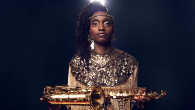 Jazz saxophonist Lakecia Benjamin, a young Black woman with twists pulled up into a side ponytail, wears a gold sequined shirt and a headpiece that looks a bit like a halo around her forehead. She holds her saxophone horizontally in both hands.