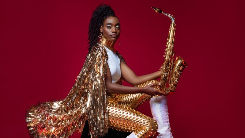 Jazz saxophonist Lakecia Benjamin, a young Black woman with twists pulled up into a side ponytail, wears a gold cape and pants and white shirt and boots. She sits in front of a red background and holds her saxophone up and away from her body, gazing unconcernedly towards the camera. 