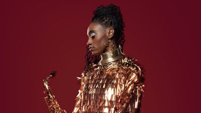 Jazz saxophonist Lakecia Benjamin, a young Black woman with twists pulled up into a side ponytail, wears a gold cape gazes down towards her saxophone in front of a red background.