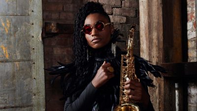 Jazz saxophonist Lakecia Benjamin, a young Black woman with twists pulled up into a side ponytail, wears a a black feathered shirt and red round glasses. She stands in front of an industrial looking wall and holds her saxophone.