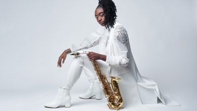 Jazz saxophonist Lakecia Benjamin, a young Black woman with twists pulled up into a side ponytail, wears all white and sits on a white block in front of a white background. She holds her saxophone by the neck and rests it on the floor next to her.