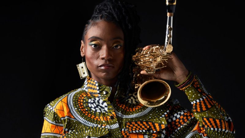 Jazz saxophonist Lakecia Benjamin, a young Black woman with twists pulled up into a side ponytail, wears an orange and green patterned jumpsuit and holds rests her saxophone on her shoulder.