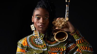 Jazz saxophonist Lakecia Benjamin, a young Black woman with twists pulled up into a side ponytail, wears an orange and green patterned jumpsuit and holds rests her saxophone on her shoulder.