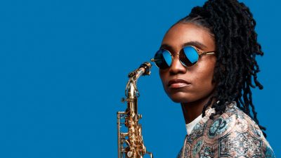 Jazz saxophonist Lakecia Benjamin, a young Black woman with twists pulled up into a side ponytail, wears a blue and pink patterned satin shirt and blue round glasses. She holds her saxophone up near her face in front of a stark cornflower blue background.