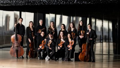 The members of Les Arts Florissants and violinist Theotime Langlois de Swarte wear black concert attire and pose with their instruments in a room with a wall of windows, a wall of mirrors, hardwood floors, and a dark but shiny ceiling.