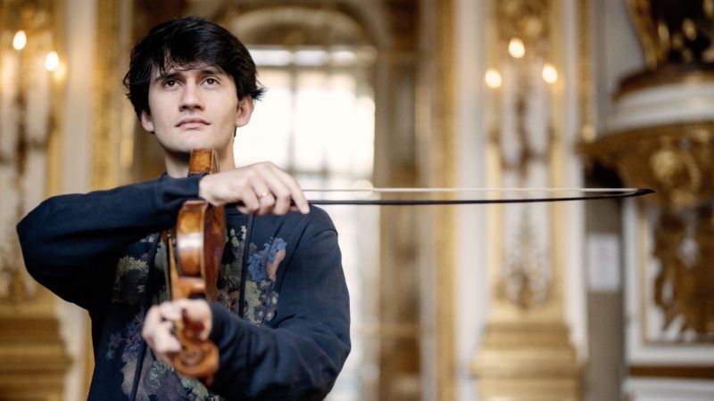 Violinist Theotime Langlois de Swarte, a young white man with medium length straight dark brown hair, wears a navy blue patterned button down shirt in an ornate hallway. He holds his violin sideways and holds his bow horizontally, perpendicular to the violin, across the frame.