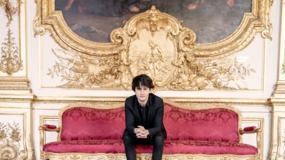 Violinist Theotime Langlois de Swarte, a young white man with medium length straight dark brown hair, wears a black suit in an ornate hallway, sitting on a red couch in front of an old painting with a very ornate gold frame.