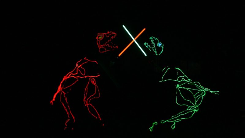 Light art T-rexs duel with lightsabers in a scene from Lightwire Theater's "Dino-Light."