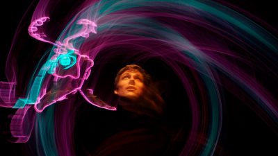 A member of Lightwire Theater stands in a swirl of purple and blue light art, a fish near his face, all against a black background.