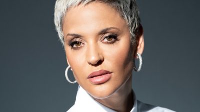 Fado singer Mariza, a Portuguese woman with short bleached blonde hair, brown eyes, silver hoops, and dusty rose lips, looks towards the camera in front of a dark background.