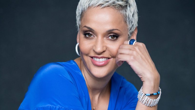 Fado singer Mariza, a Portuguese woman with short bleached blonde hair, brown eyes, silver hoops,a sapphire ring, and dusty rose lips, wears a royal blue blouse and looks towards the camera in front of a dark background.