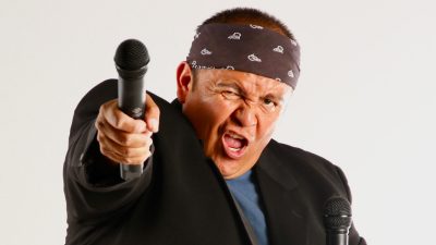 Indigenous commedian Ernest Tsosie, a Native middle aged man in a blue T-shirt and black blazer with a black and white patterned bandana tied around his head, winks one eye and holds a microphone towards the camera.