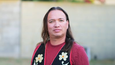 Comedian Jim Ruel, a middle aged Indigenous man with long straight brown hair, wears a red long-sleeved T-shirt under a black floral patterned vest and looks towards the camera. The photo was taken outside.