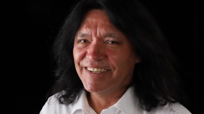 Indigenous comedian Marc Yaffee, a middle aged Native man with shoulder-length dark brown hair, wears a white button down shirt in front of a black background and smiles towards the camera.