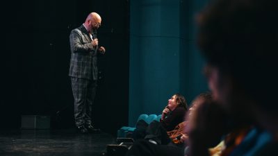 A bald white man in a grey plaid suit emcees Ontroerend Goed's "Fight Night" and speaks to an audience member in the front row.