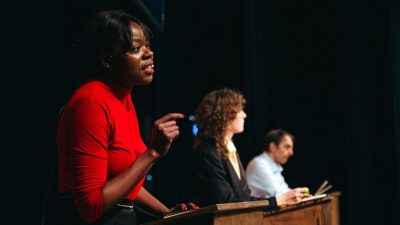 Some of the cast of Ontroerend Goed's "Fight Night" perform on stage; three people, one Black woman, one white woman, and one white man, speak from podiums as if they're in a presidential debate.