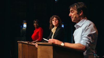 Some of the cast of Ontroerend Goed's "Fight Night" perform on stage; three people, one Black woman, one white woman, and one white man, speak from podiums as if they're in a presidential debate.