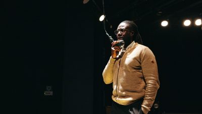 A cast member of Ontroerend Goed's "Fight Night," a Black man with dreds pulled back into a ponytail wearing a beige sweater, speaks into a hanging microphone and is lit starkly from above.