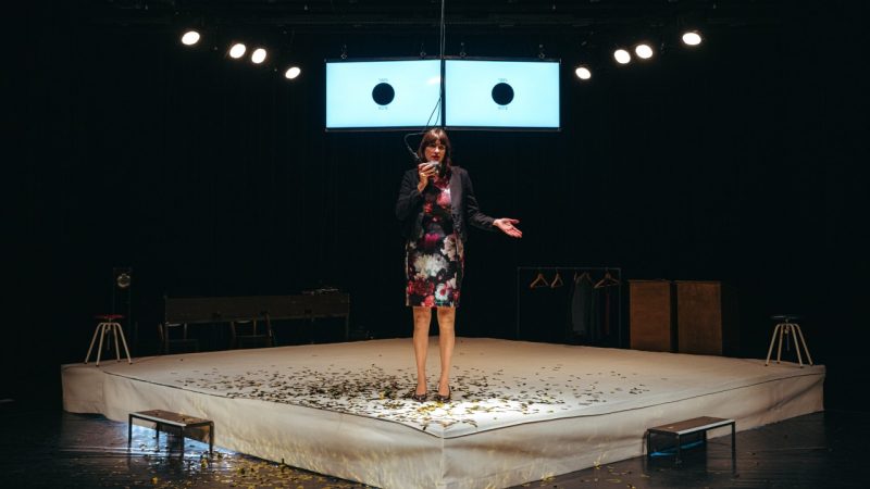 A cast member of Ontroerend Goed's "Fight Night," a white woman wearing a patterned floral dress and dark grey blazer, speaks into a hanging microphone and is lit starkly from above. On stage at her feet is gold confetti and two screens behind her show a pie chart at 100%.