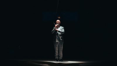 A cast member of Ontroerend Goed's "Fight Night," a white bald man wearing a plaid suit speaks into a hanging microphone and is lit starkly from above.