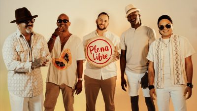 The members of Plena Libre, five Hispanic men, wear white and nude shades and stand in front of a beige wall lit from below with pale amber light.