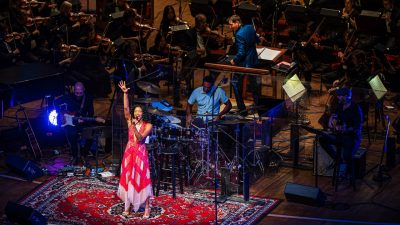 Broadway star Renée Elise Goldsberry, a young Black woman, belts out a note on stage in a long pink dress, her arm in the air above her head, as an orchestra performs behind her.