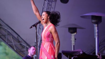 Broadway star Renée Elise Goldsberry, a young Black woman, belts out a note on stage in a long pink patterned dress, her arm in the air above her head.