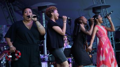 Broadway star Renée Elise Goldsberry, a young Black woman, belts out a note on stage in a long pink patterned dress, along with her three backup singers, Black women in black dresses.
