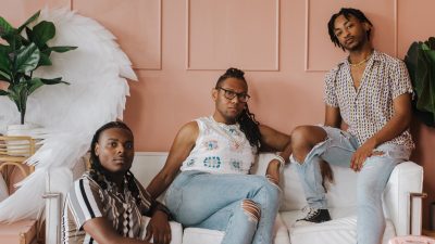 The members of the Harlem Gospel Travelers, three young Black singers wearing casual clothes and sitting or leaning on a white couch in a pink room.