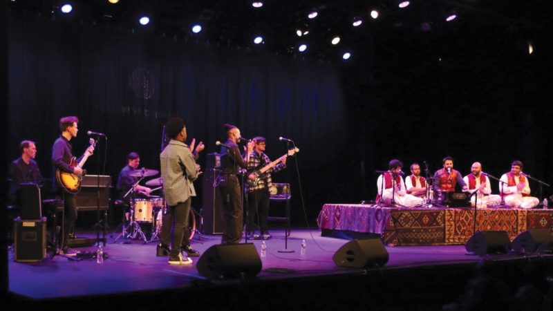 Members of Riyaaz Qawwali and the Harlem Gospel Travelers perform together on stage under purple light. The Riyaaz Qawwali members sit on a raised platform draped in carpets.