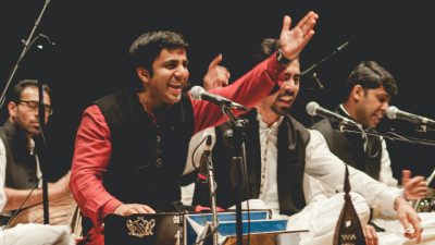The members of Riyaaz Qawwali, four brown men, perform on stage with energy, the one in the foreground raising his arm towards the crowd and a big smile on his face.