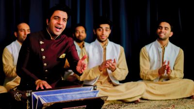 The members of Riyaaz Qawwali, five brown men, wear traditional garb and sit cross-legged as they perform on stage, the one in the foreground raising his arm gently towards the audience.