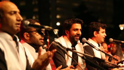 The members of Riyaaz Qawwali, four brown men, wear traditional garb as they perform on stage, all singing into microphones.