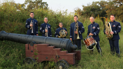 Members of the United States Air Force Heritage Brass wear blue uniforms and hold their instruments in a grass field next to a cannon.