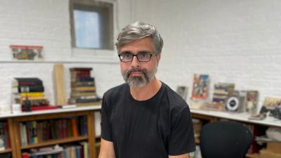Artist Brian Dettmer, a middle aged white man wearing a black T-shirt and black framed glasses. His hair and beard are salt and pepper, and he is surrounded by books, materials, and completed works in his art studio.