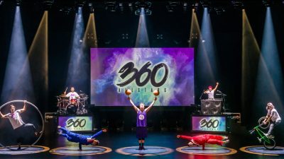 Members of 360 ALLSTARS perform on stage, two break dance, one on a BMX bike, one in a German wheel, another balancing two basketballs on the tips of his fingers.