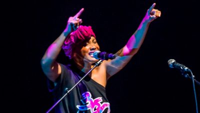 Members of 360 ALLSTARS perform on stage, a young Black woman in a pink head wrap DJing and singing animatedly into a microphone.