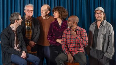 Bill Frisell, second from left, with musical collaborators (from left) Thomas Morgan, Hank Roberts, Jenny Scheinman, Rudy Royston, and Eyvind Kang. They stand in casual clothes in front of a rich teal curtain.