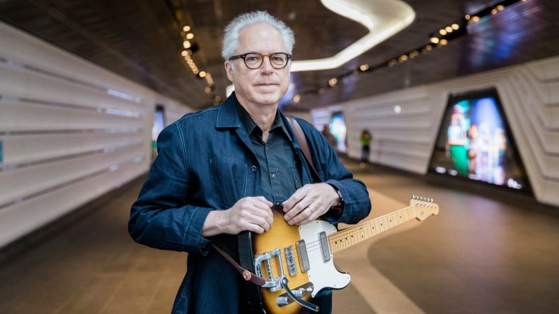 Guitarist Bill Frisell, a white man with white hair and tortoise shell glasses, stands in a futuristic hallway, his electric guitar slung around his neck, in a denim button down shirt.
