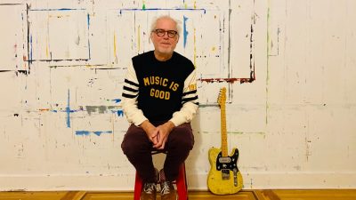 Jazz guitarist Bill Frisell, a white man with dark framed glasses and white hair, sits on a red stool near a yellow electric guitar. The wall behind him has outlines of previous paintings that were created while hanging on the wall.